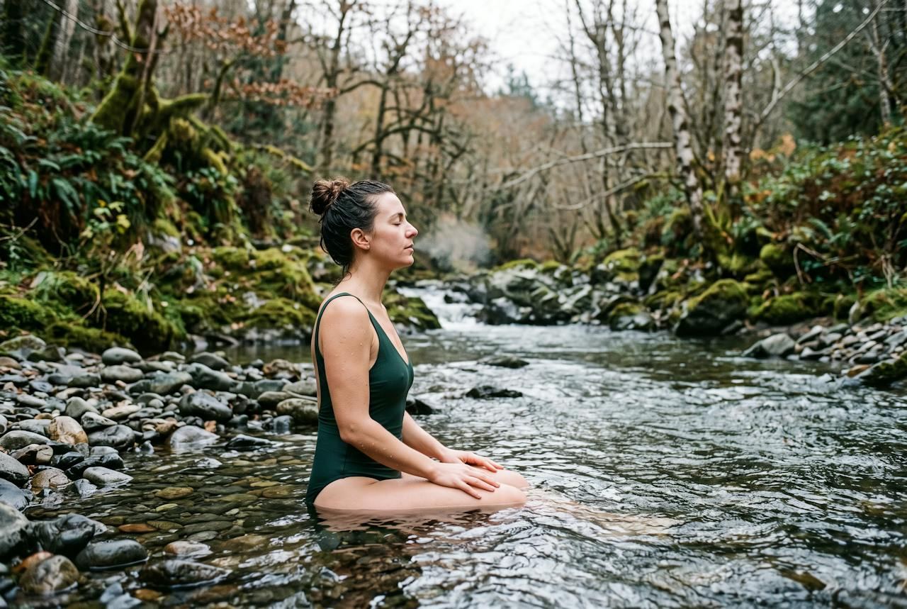 A woman sitting in a cold forest creek with her eyes closed.